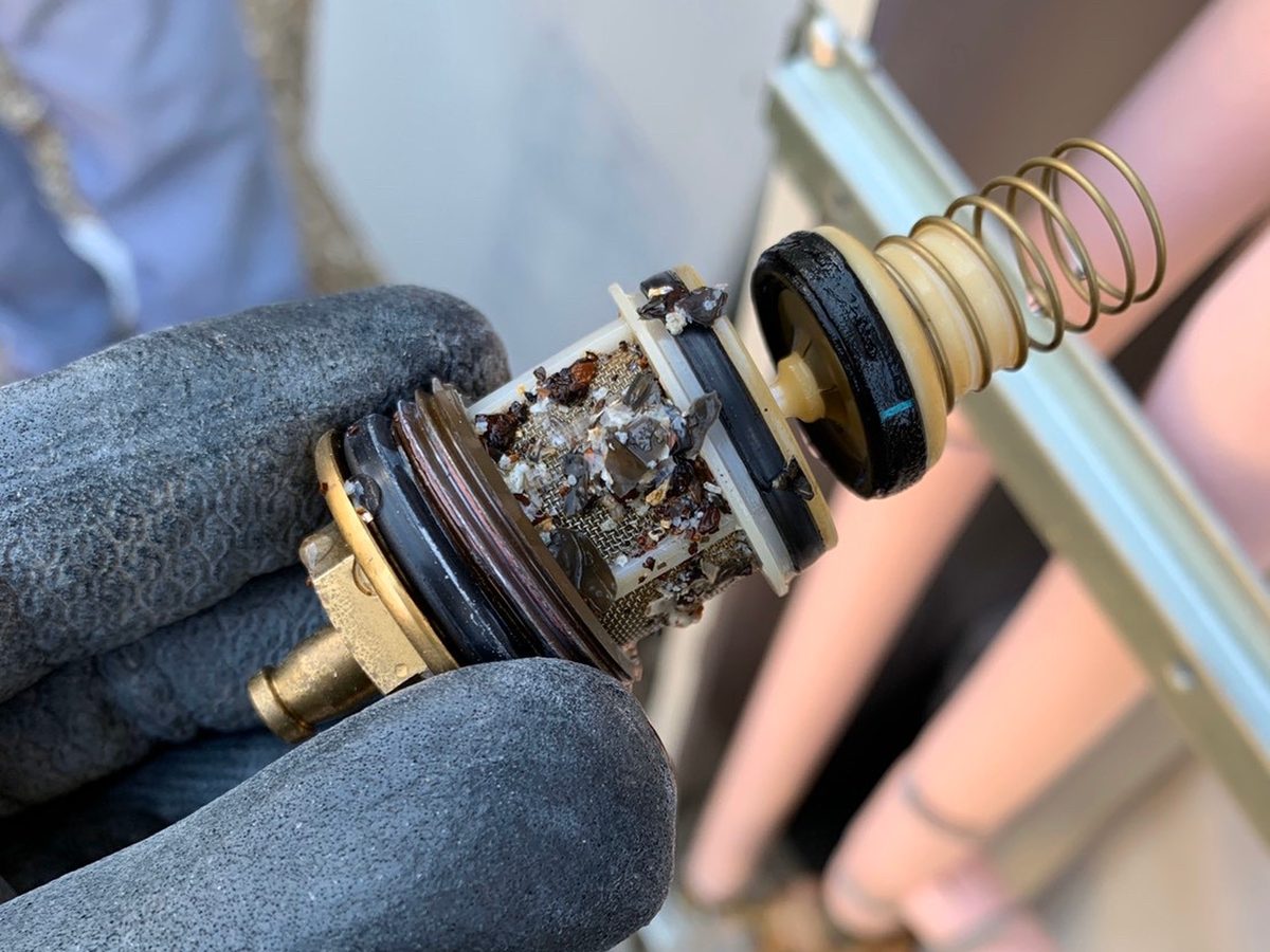 作業手袋で持った給湯配管のストレーナ内部。金属の網目に赤茶色のサビ片と白いスケールがびっしり詰まっている接写