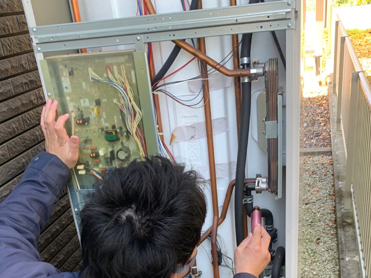 作業着の技術者が屋外機のパネルを外し、制御基板と銅配管が見える状態で点検している様子